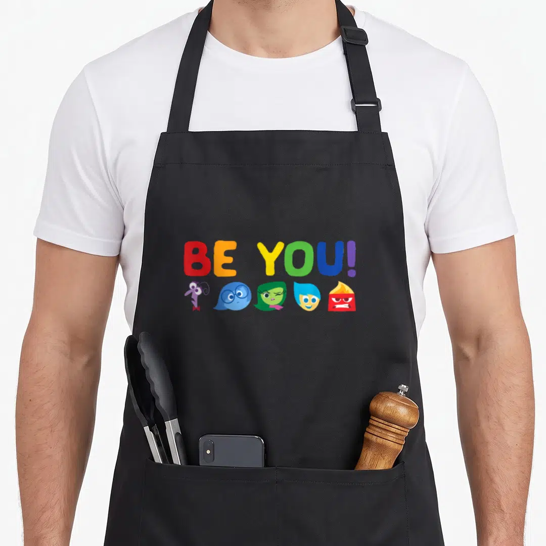 Cool rainbow apron - BE YOU! text in rainbow colors above five cartoon character heads — shown on a black cotton canvas apron against a white background