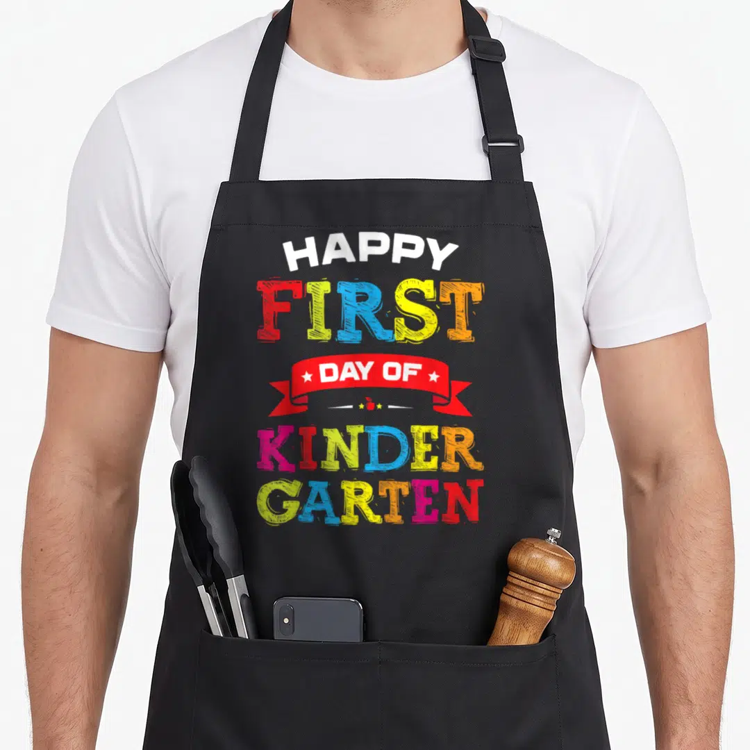 Kindergarten art teacher apron - Stacked colorful text reading Happy First Day of Kindergarten with red ribbon and tiny — shown on a black cotton canvas apron against a white background