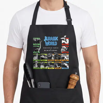 Jurassic table apron - Periodic table of dinosaurs chart with 14 species illustrations and colored borders — shown on a black cotton canvas apron against a white background