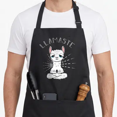 Llamaste grill instructor apron - Cartoon llama meditating in lotus pose with LLAMASTE text and white rays — shown on a black cotton canvas apron against a white background
