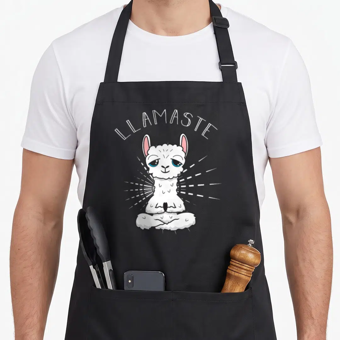 Llamaste grill instructor apron - Cartoon llama meditating in lotus pose with LLAMASTE text and white rays — shown on a black cotton canvas apron against a white background