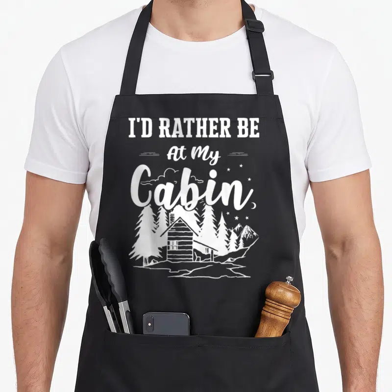 Rather cabin apron - White text I'D RATHER BE At My Cabin above cabin, pines, and stars night scene — shown on a black cotton canvas apron against a white background