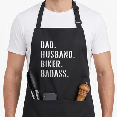 Biker wife apron - White distressed text DAD HUSBAND BIKER BADASS on black background — shown on a black cotton canvas apron against a white background
