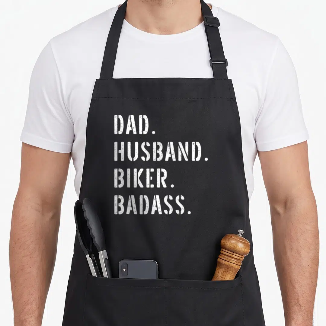 Biker wife apron - White distressed text DAD HUSBAND BIKER BADASS on black background — shown on a black cotton canvas apron against a white background