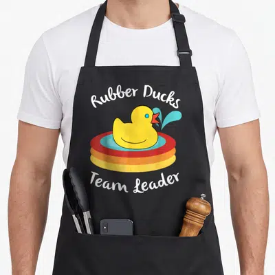 Rubber water play aprons - Cartoon yellow duck in red and blue float with Rubber Ducks Team Leader text — shown on a black cotton canvas apron against a white background