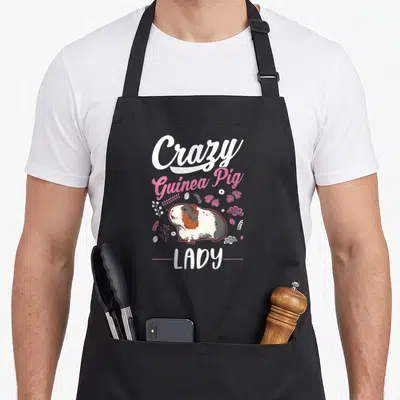 Retro animal crazy guinea lady apron - Cartoon guinea pig with white pink brown fur and floral accents — shown on a black cotton canvas apron against a white background