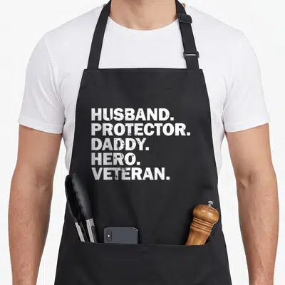 Protector hero best husband apron - White distressed text stack reading HUSBAND PROTECTOR DADDY HERO VETERAN on black — shown on a black cotton canvas apron against a white background