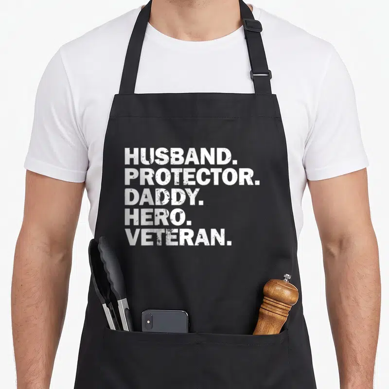 Protector hero best husband apron - White distressed text stack reading HUSBAND PROTECTOR DADDY HERO VETERAN on black — shown on a black cotton canvas apron against a white background