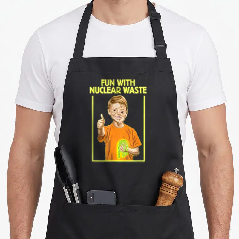 Nuclear funniest aprons - Cartoon boy in orange shirt holding glowing green slime under yellow text — shown on a black cotton canvas apron against a white background