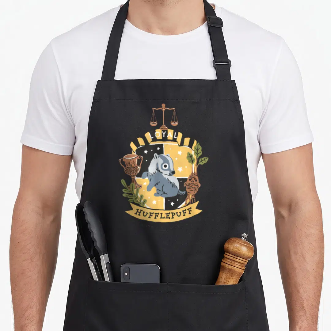 Deathly hufflepuff apron - Cartoon badger shield with scales, cup, root, and text LOYAL HUFFLEPUFF DEDICATION PATIENCE — shown on a black cotton canvas apron against a white background
