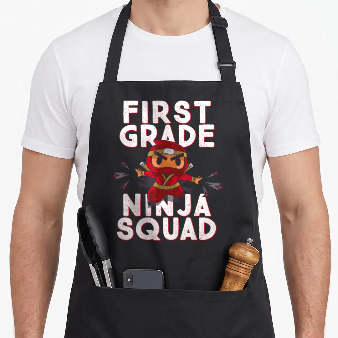 1st grade squad apron - Cartoon ninja surrounded by six shuriken with bold white and red text on grey — shown on a black cotton canvas apron against a white background