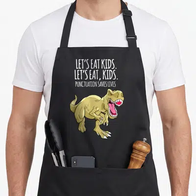 Live grammar let's eat apron - Cartoon T-Rex below stacked text reading Let's Eat Kids Punctuation Saves Lives — shown on a black cotton canvas apron against a white background