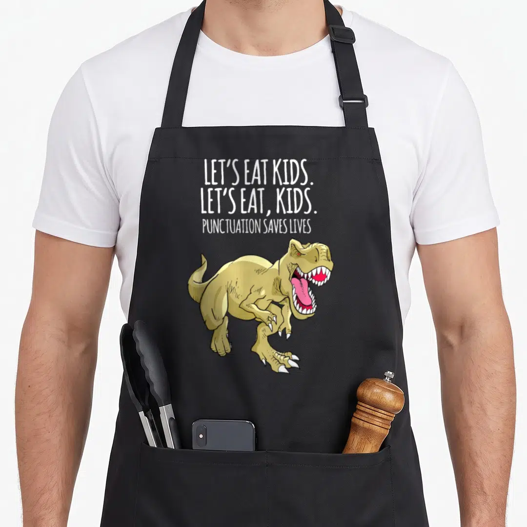 Live grammar let's eat apron - Cartoon T-Rex below stacked text reading Let's Eat Kids Punctuation Saves Lives — shown on a black cotton canvas apron against a white background