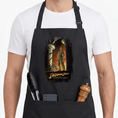 Doom temple apron - Indiana Jones standing in temple doorway with vines and stacked movie title text in brown and yellow — shown on a black cotton canvas apron against a white background