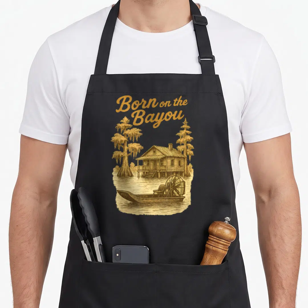 Louisiana southern aprons - Gold Born on the Bayou text above airboat, cypress trees, and stilt house scene — shown on a black cotton canvas apron against a white background