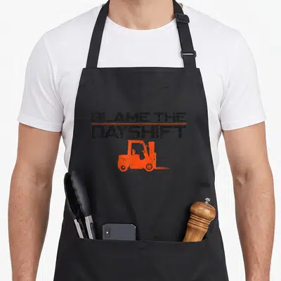Certified truck apron - Distressed BLAME THE DAYSHIFT text above orange forklift silhouette on grey background — shown on a black cotton canvas apron against a white background