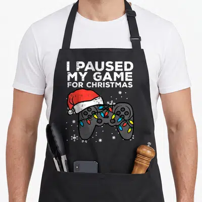Paused christmas apron - I PAUSED MY GAME FOR CHRISTMAS text above grey controller with Santa hat and colorful lights — shown on a black cotton canvas apron against a white background