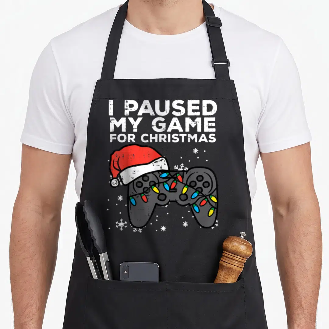 Paused christmas apron - I PAUSED MY GAME FOR CHRISTMAS text above grey controller with Santa hat and colorful lights — shown on a black cotton canvas apron against a white background