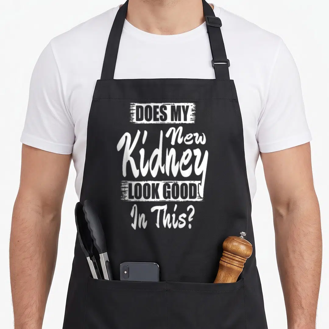 Transplant recipient organ apron - White stacked typography reading DOES MY New Kidney LOOK GOOD In This? on gray — shown on a black cotton canvas apron against a white background