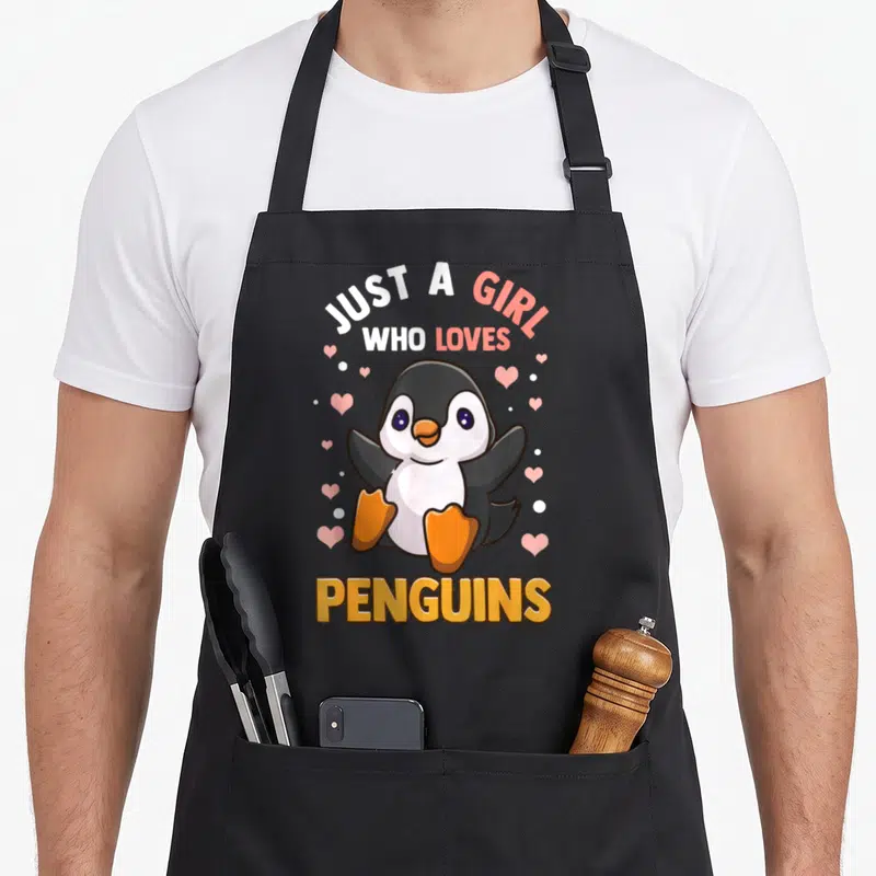 Vintage penguin aprons - Cartoon penguin with pink hearts and text Just A Girl Who Loves Penguins — shown on a black cotton canvas apron against a white background