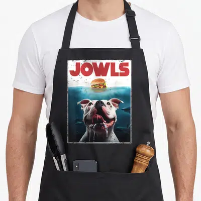 Bulldog jowl dog apron - JOWLS text above submerged bulldog eyeing floating burger in blue water with sharks — shown on a black cotton canvas apron against a white background