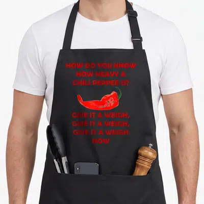 Chili heavy apron - Red chili pepper between red text reading How Do You Know How Heavy A Chili Pepper Is Give It — shown on a black cotton canvas apron against a white background