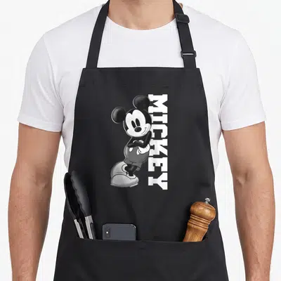 Funny lean best friend aprons - Mickey Mouse leaning against vertical white text reading MICKEY in black and white — shown on a black cotton canvas apron against a white background