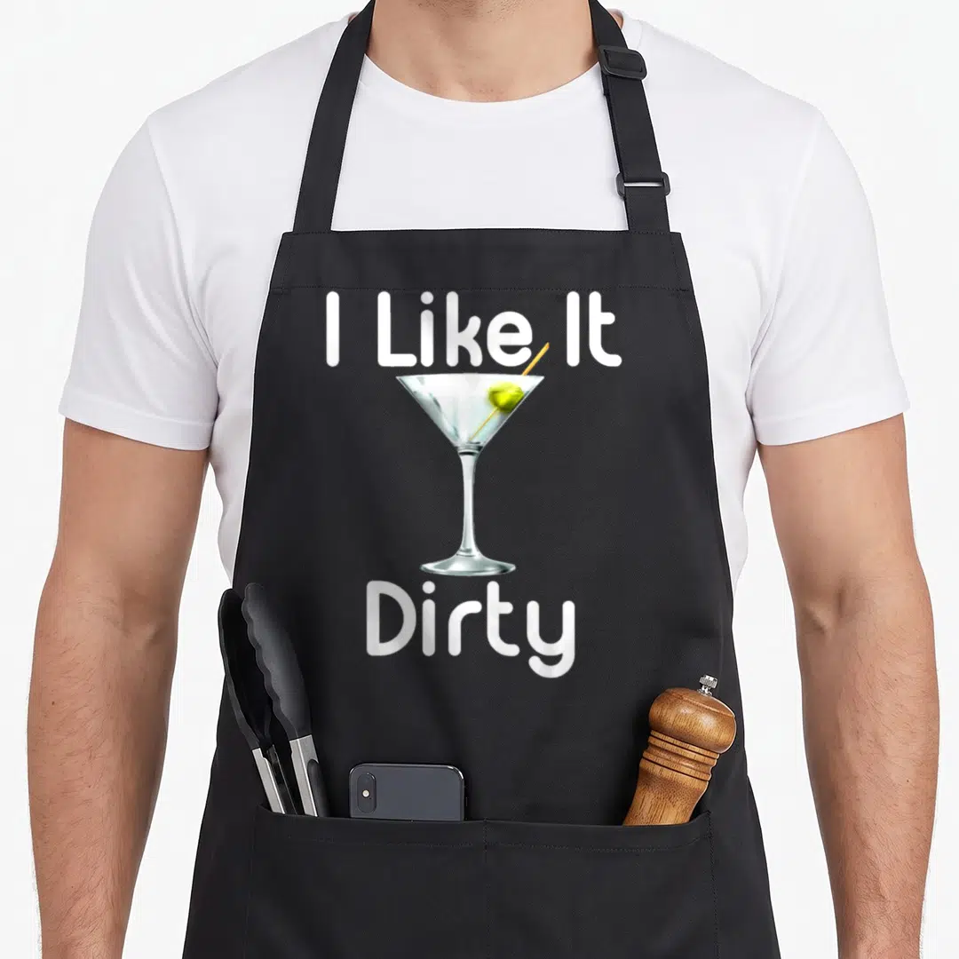 Hour the dirty apron - I Like It Dirty text framing a realistic martini glass with green olive — shown on a black cotton canvas apron against a white background
