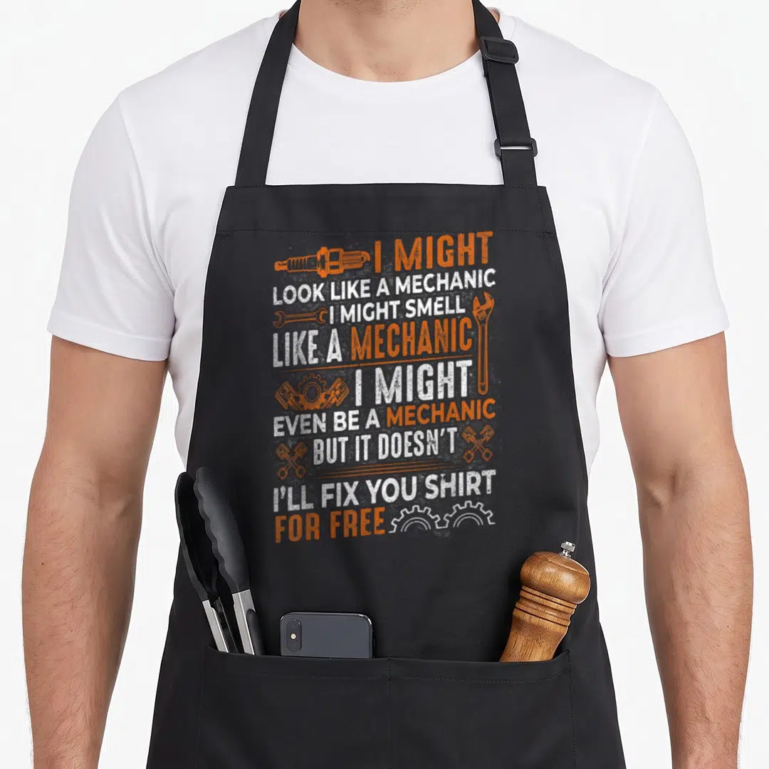 Might auto mechanic apron - Stacked orange and white text reading I might look like a mechanic with spark plug — shown on a black cotton canvas apron against a white background