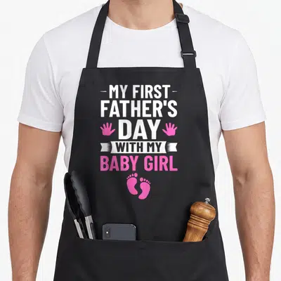 Outfit father day apron - My First Fathers Day with my baby girl text, pink handprints, and footprints — shown on a black cotton canvas apron against a white background
