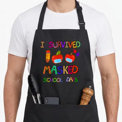Survived school apron - I Survived Masked School Days text with cartoon apples wearing masks and handprints — shown on a black cotton canvas apron against a white background