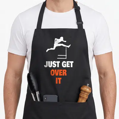 Field jumping track apron - White hurdler silhouette jumping over a hurdle above stacked text JUST GET OVER IT in white — shown on a black cotton canvas apron against a white background