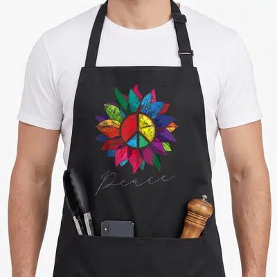 Distressed sunflower apron - multicolored sunflower with peace symbol and cursive Peace text — shown on a black cotton canvas apron against a white background