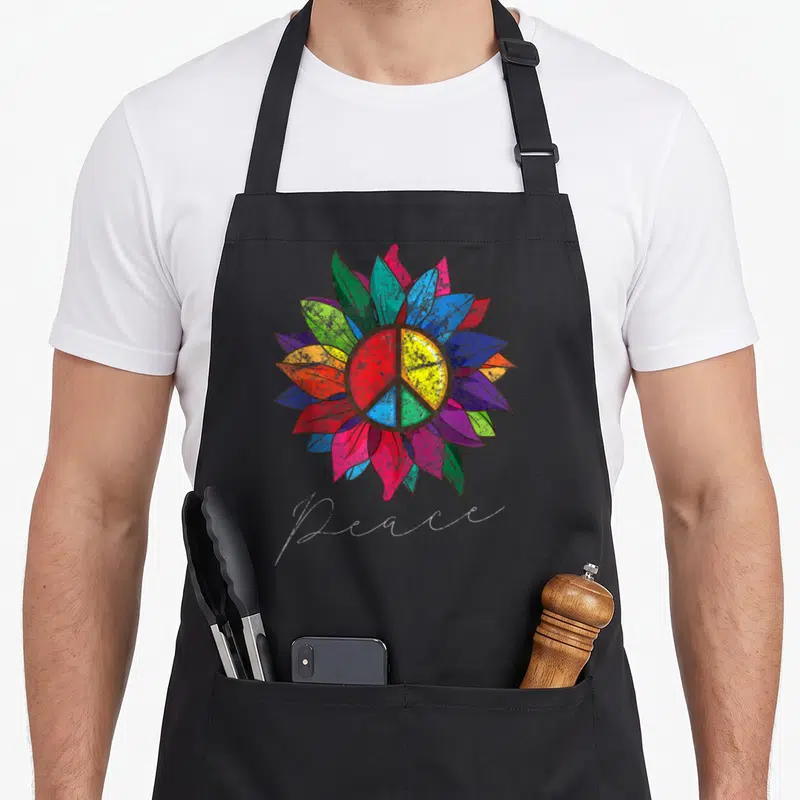 Distressed sunflower apron - multicolored sunflower with peace symbol and cursive Peace text — shown on a black cotton canvas apron against a white background
