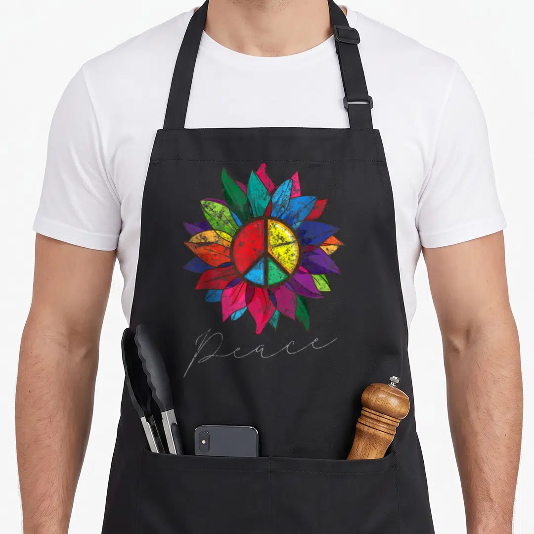 Distressed sunflower apron - multicolored sunflower with peace symbol and cursive Peace text — shown on a black cotton canvas apron against a white background