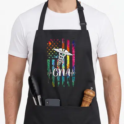 Flag tie-dye apron - White caduceus over tie-dye flag with CNA script and heartbeat line — shown on a black cotton canvas apron against a white background