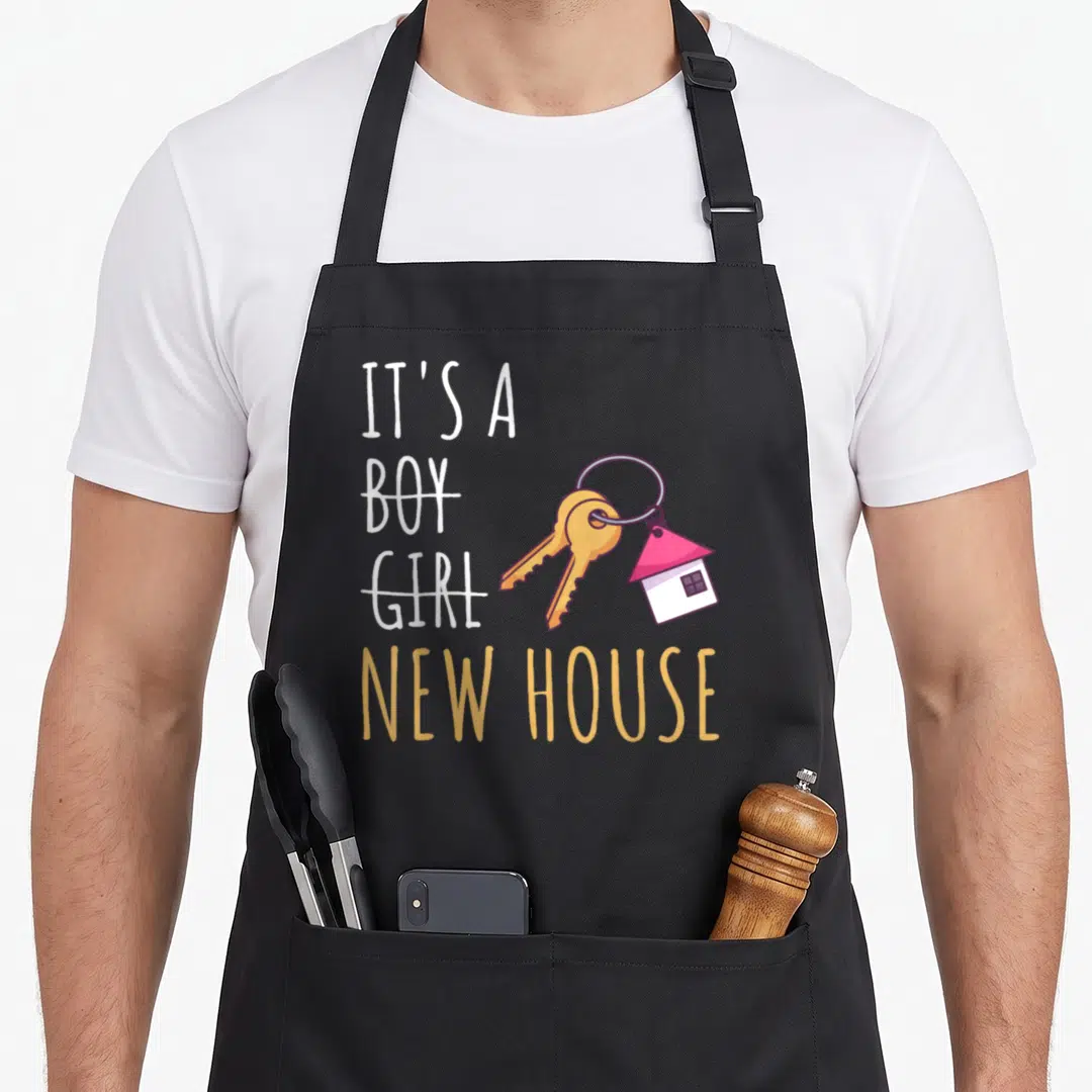 Owner housewarming apron - Text it's a new house with crossed out boy girl and golden keys with pink house charm — shown on a black cotton canvas apron against a white background