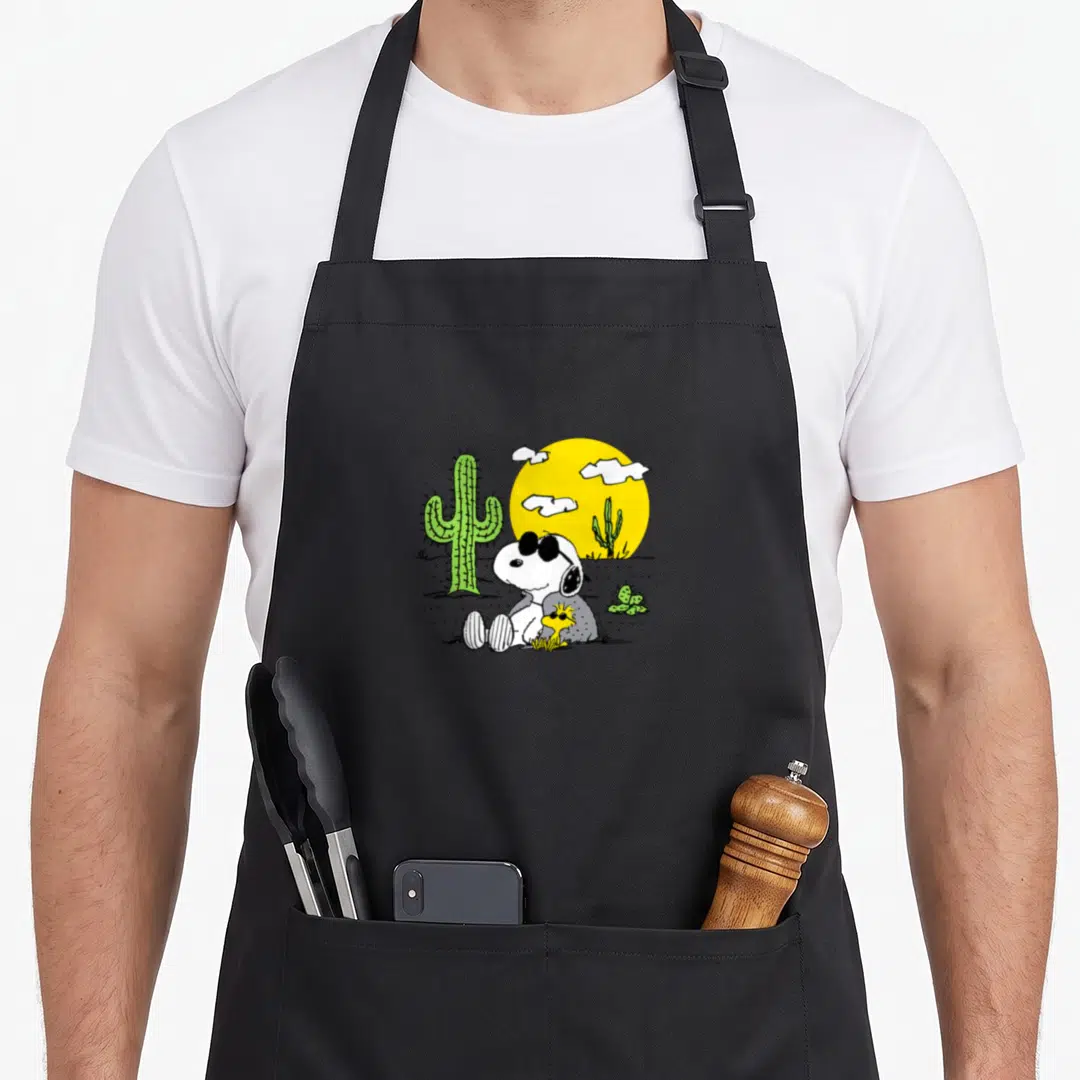 Woodstock summer aprons - Snoopy and Woodstock sitting in a desert with a large green cactus and yellow sun — shown on a black cotton canvas apron against a white background