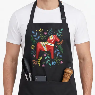 Swedish dala horse apron - Red cartoon Dala horse surrounded by blue yellow green and pink leaves and flowers — shown on a black cotton canvas apron against a white background