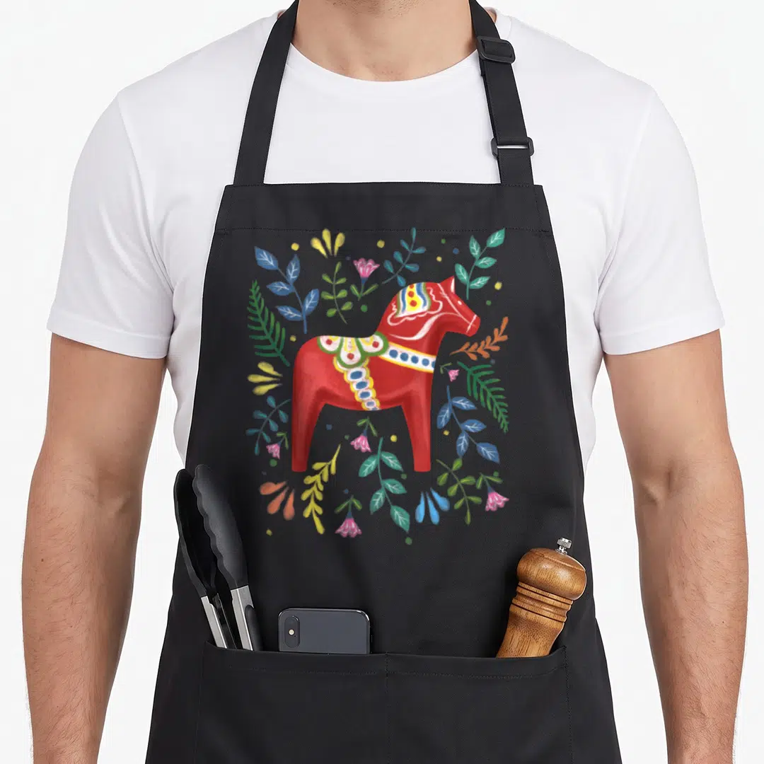 Swedish dala horse apron - Red cartoon Dala horse surrounded by blue yellow green and pink leaves and flowers — shown on a black cotton canvas apron against a white background