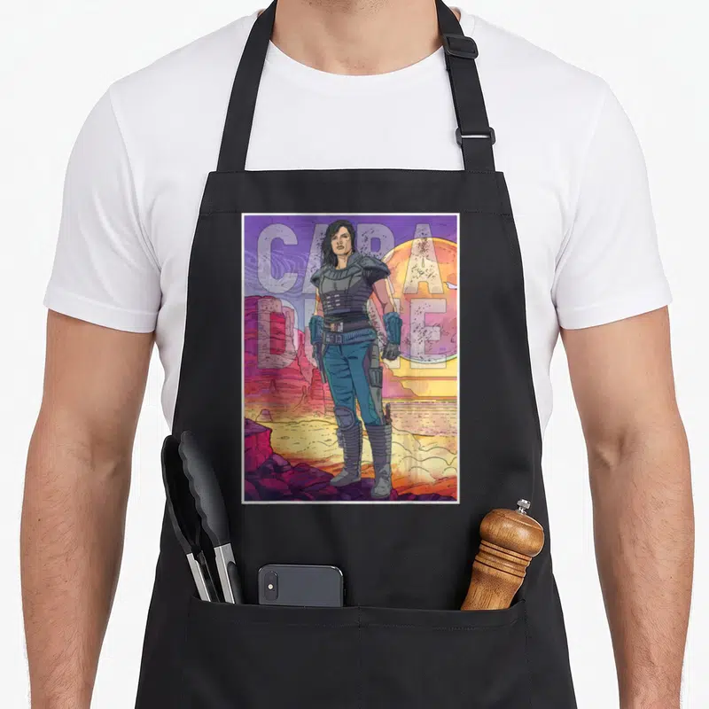 Cara dunes apron - Cartoon Cara Dune standing in purple and orange desert with large split text — shown on a black cotton canvas apron against a white background