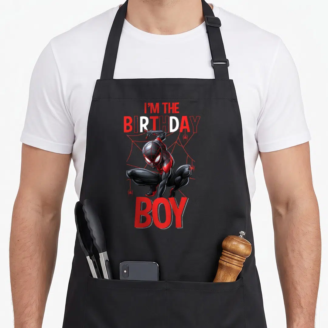 Funny mile birthday apron - Spider-Man crouching below I'M THE BIRTHDAY BOY text over red web background — shown on a black cotton canvas apron against a white background