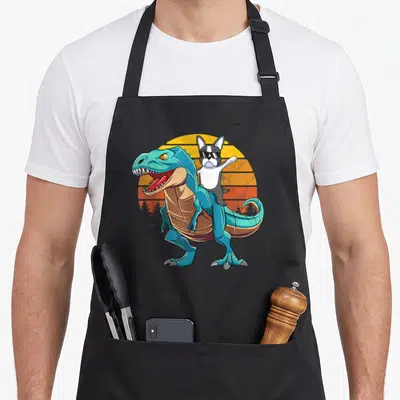 Riding dog apron - Cartoon Boston Terrier wearing sunglasses riding a blue dinosaur against a striped retro sunset — shown on a black cotton canvas apron against a white background
