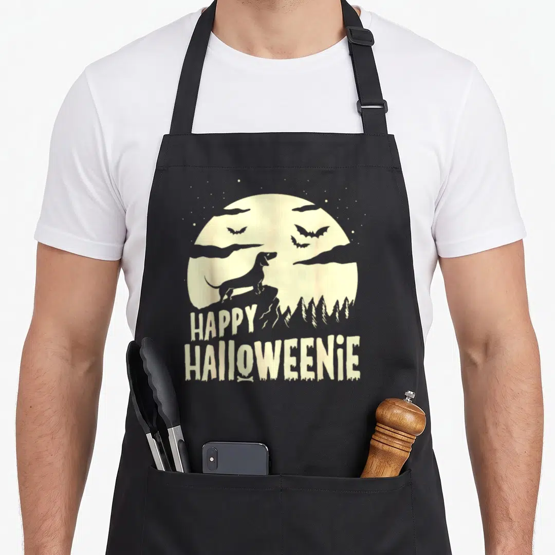 Weiner cheap halloween aprons - Dachshund silhouette on a yellow moon with stacked HAPPY HALLOWEENIE text and a bone — shown on a black cotton canvas apron against a white background