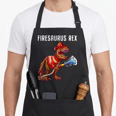 Rex firefighter apron - Cartoon Tyrannosaurus Rex in red helmet and gear holding a hose spraying blue water under white — shown on a black cotton canvas apron against a white background
