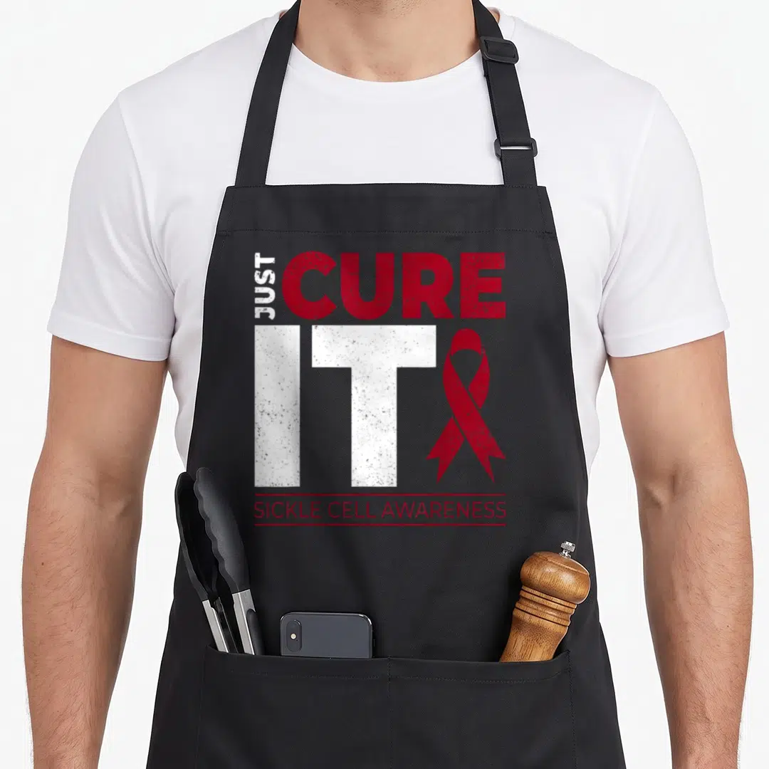 Scd ribbon apron - Distressed red and white Just Cure It text with sickle cell awareness ribbon — shown on a black cotton canvas apron against a white background
