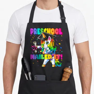 Flossing preschool apron - Rainbow unicorn in graduation cap holding diploma with text PRESCHOOL NAILED IT! — shown on a black cotton canvas apron against a white background