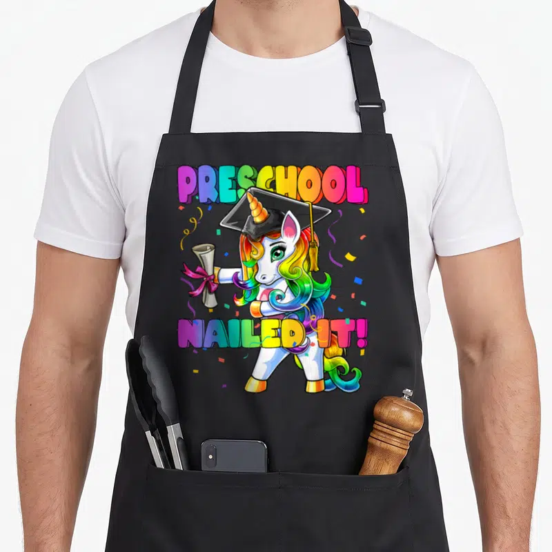 Flossing preschool apron - Rainbow unicorn in graduation cap holding diploma with text PRESCHOOL NAILED IT! — shown on a black cotton canvas apron against a white background