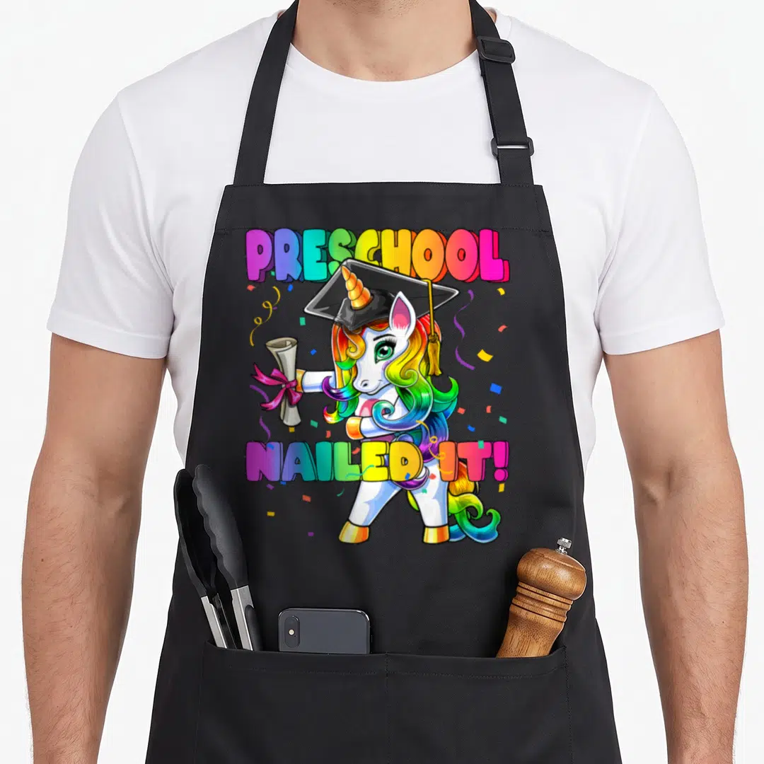 Flossing preschool apron - Rainbow unicorn in graduation cap holding diploma with text PRESCHOOL NAILED IT! — shown on a black cotton canvas apron against a white background