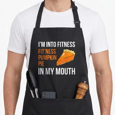 Fitness thanksgiving day aprons - Cartoon pumpkin pie slice next to white and orange text reading I'M INTO FITNESS — shown on a black cotton canvas apron against a white background